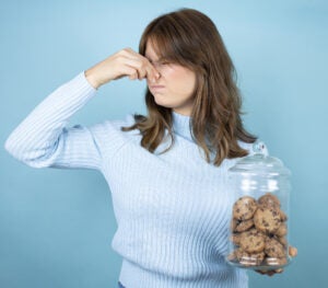 Young beautiful woman holding chocolate chips cookies jar over isolated blue background smelling something stinky and disgusting, intolerable smell, holding breath with fingers on nose