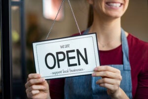 Small business owner smiling while turning the sign for the reopening of the place after the quarantine due to covid-19. Close up of woman’s hands holding sign now we are open support local business.